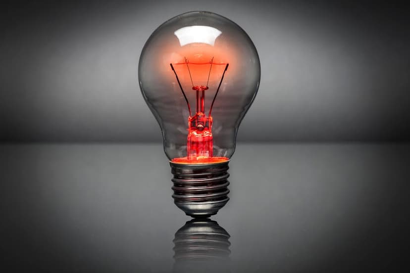 Clear incandescent bulb with a glowing red filament rests on a reflective dark surface against a gray background.
