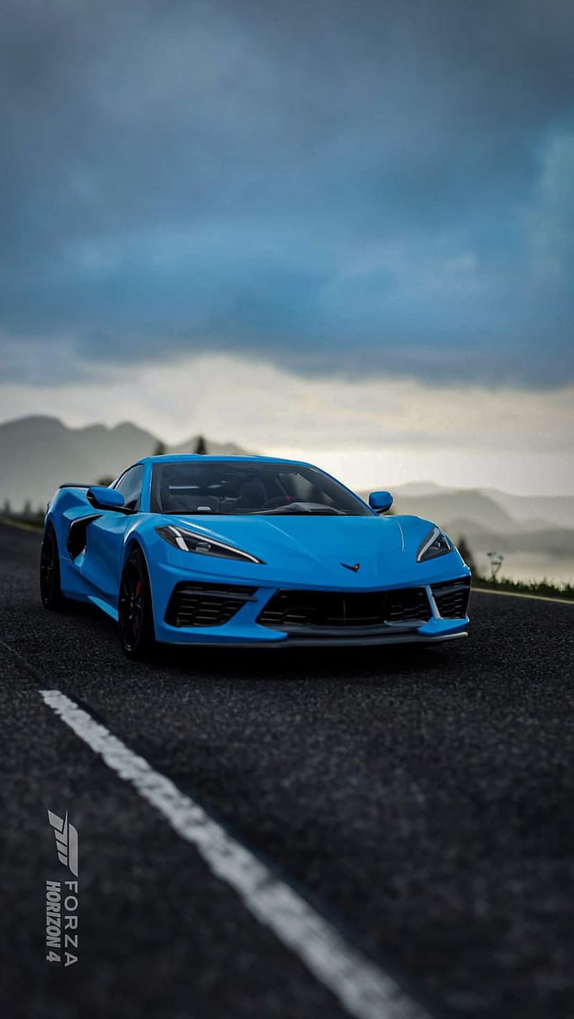 Blue Chevrolet Corvette C8 driving on a winding asphalt road with jagged gray mountains under a cloudy sky.