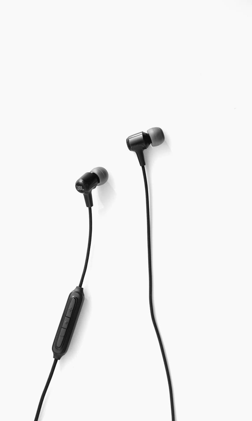 Black JBL wireless earbuds with a visible charging cable sit diagonally on a bright white studio background.