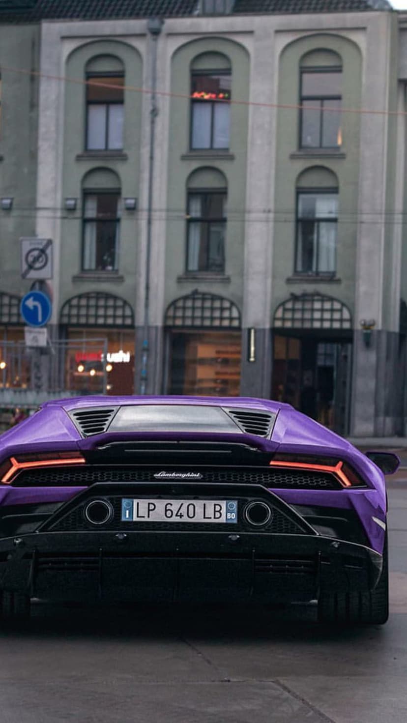 Purple Lamborghini rear view with glowing taillights parked on a city street near an arched stone building.
