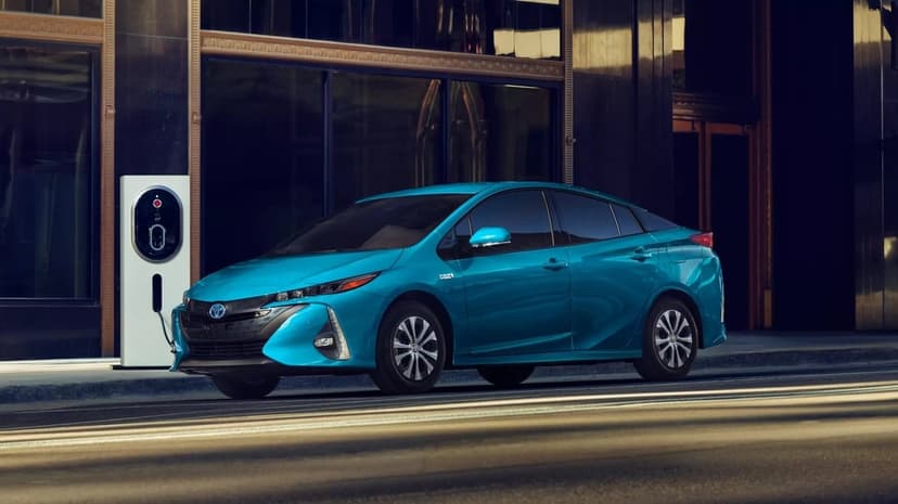 Teal blue Toyota Prius Prime parked at a white charging station in front of a dark glass building at night.