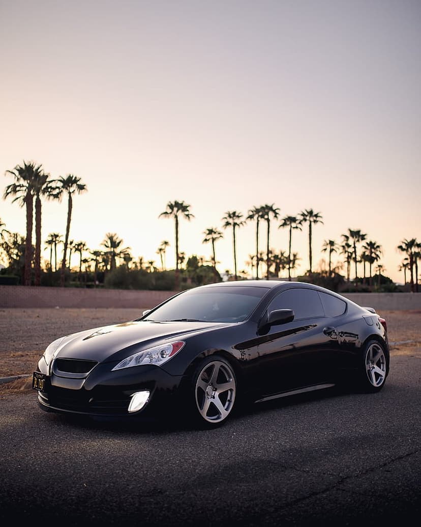 Black Hyundai Genesis Coupe parked on asphalt with palm tree silhouettes against a glowing orange sunset sky.