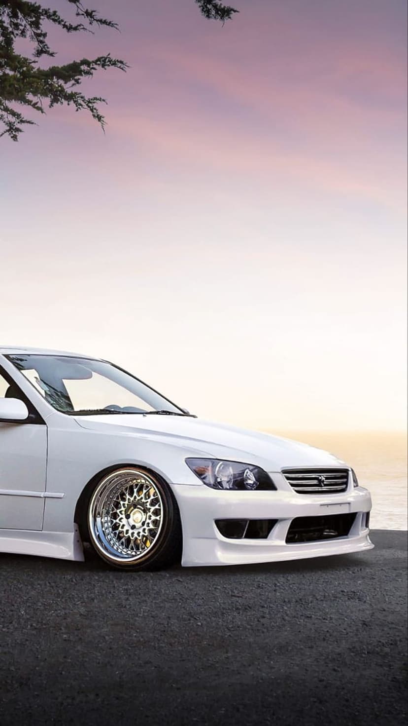 White Lexus IS sedan with lowered suspension and chrome wheels parked on a coastal road during sunset.
