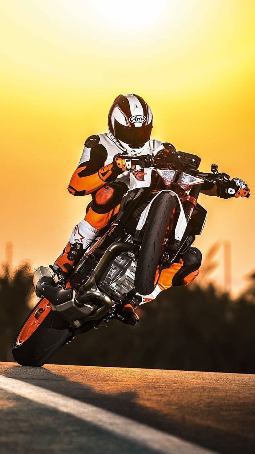Motorcyclist in a white and orange racing suit performs a high wheelie against a vivid orange sunset sky.