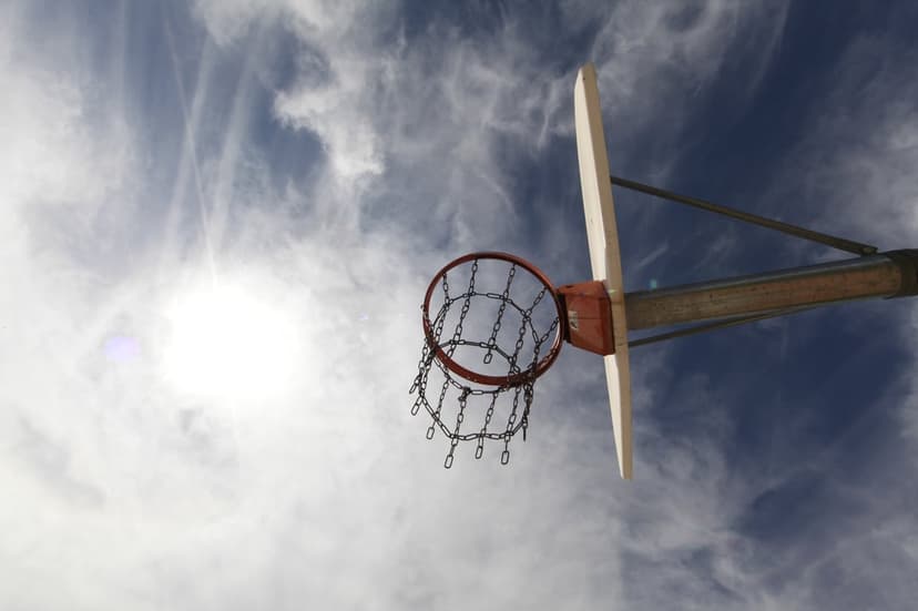 Basketball hoop with a silver chain net stands against a bright blue sky with thin clouds and a glowing sun.