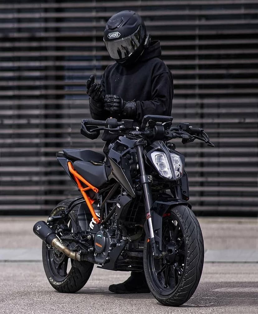 Biker in black gear stands next to a matte black sport motorcycle with orange accents against a grey wall.