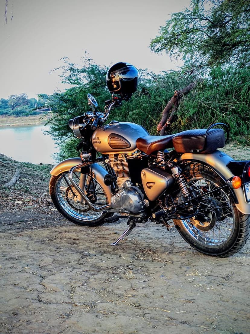 Royal Enfield motorcycle with a helmet on the handlebars parked on a dirt path by a lake during a golden sunset.