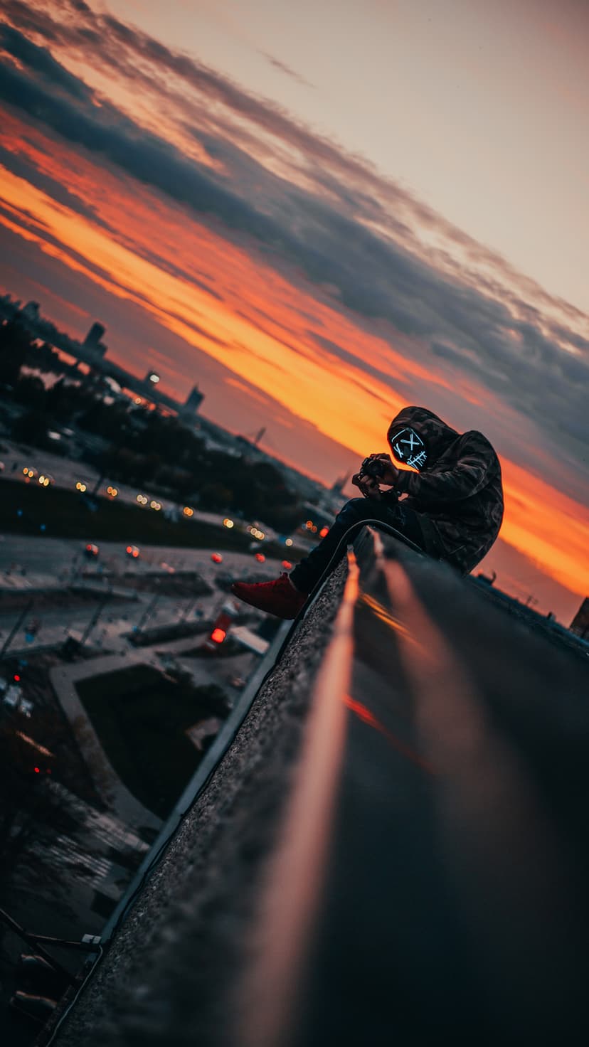 A person wearing a glowing blue X mask sits on a skyscraper ledge overlooking a fiery orange city sunset.