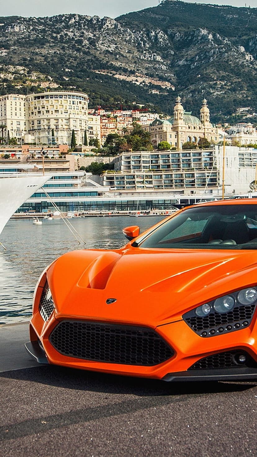 Orange supercar with sharp front grilles parked near white luxury yachts and coastal mountains in Monaco.