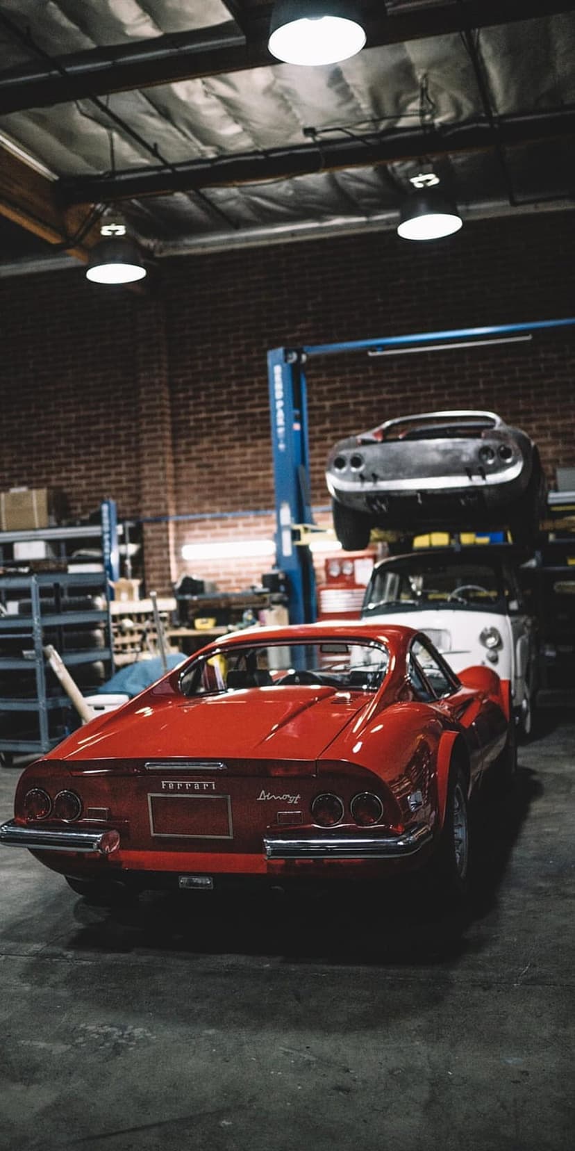 Red Ferrari Dino 246 GT parked in a brick garage under a car lift with warm industrial workshop lighting.