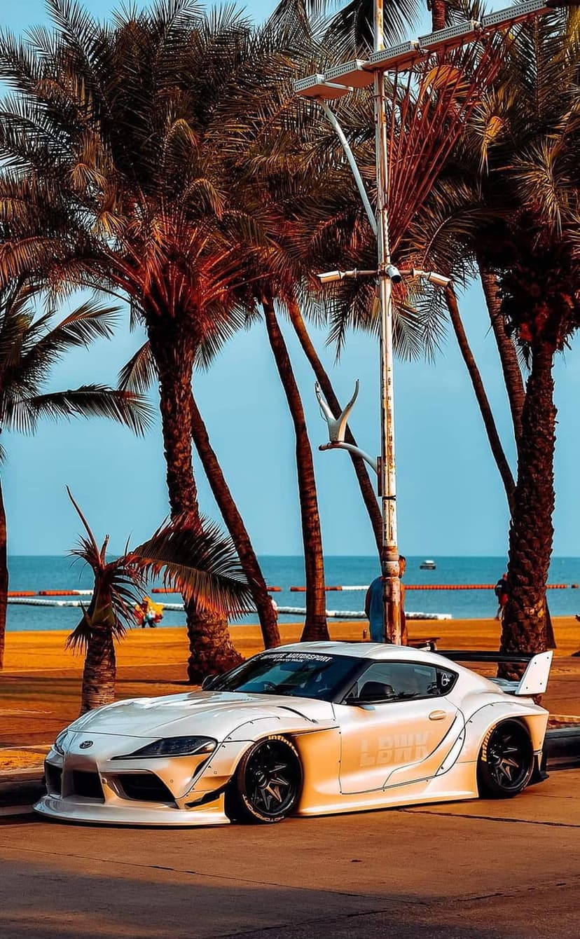White widebody Toyota Supra parked on a coastal road lined with palm trees under a clear blue sky by the ocean.