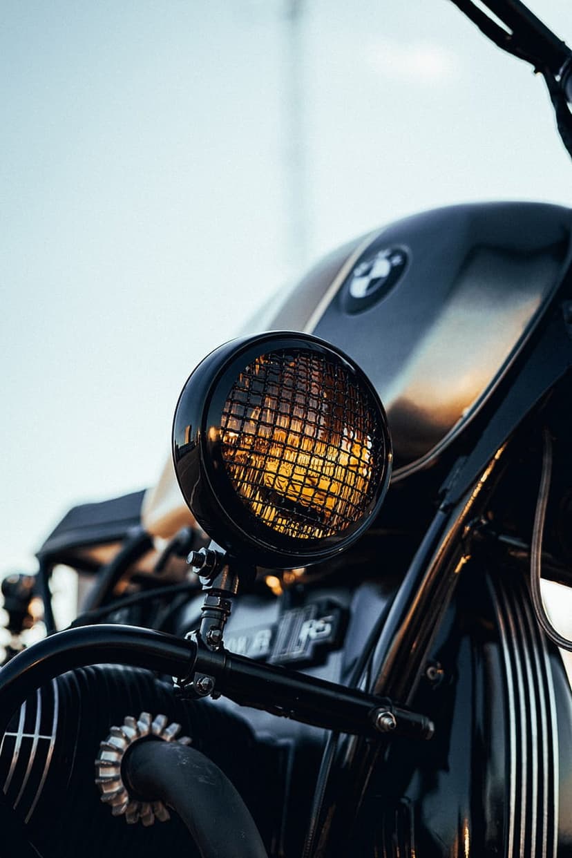 BMW motorcycle headlight with a black metal grill and warm internal glow against a bright, soft sky background.