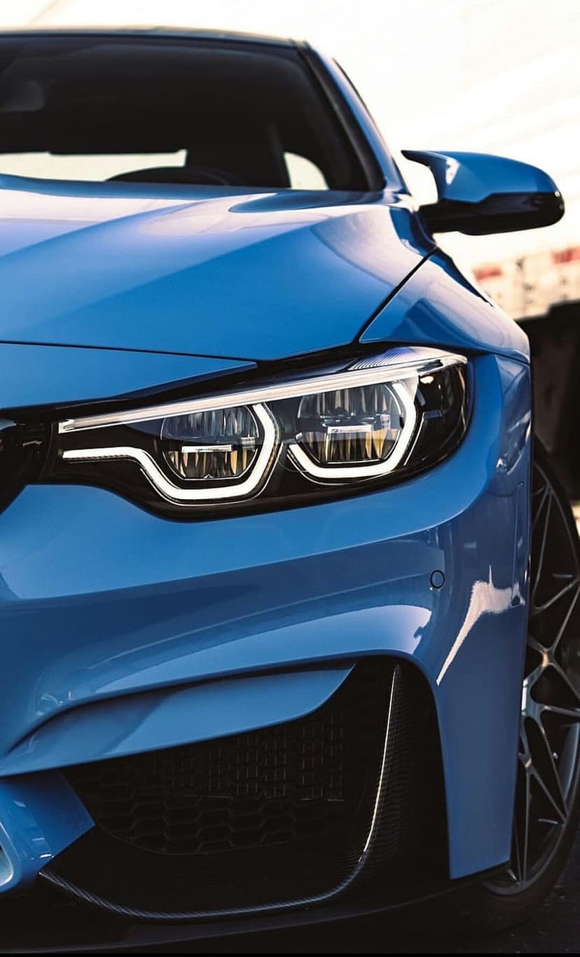 BMW M4 blue front headlight with LED daytime running lights, dark kidney grille, and carbon fiber bumper accents.