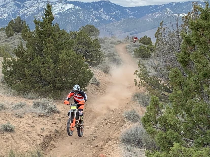 Dirt bike rider jumps mid-air on a dusty mountain trail with snow-capped peaks in the background landscape.