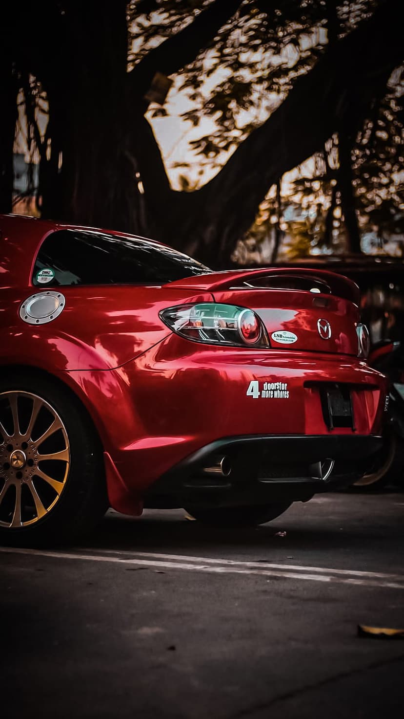 Rear view of a red Mazda RX-8 sports car with a spoiler and rotary engine badge parked near a large tree.