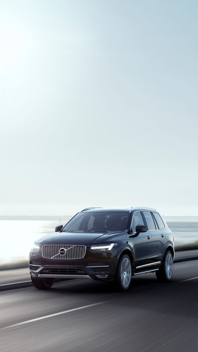 Dark Volvo XC90 SUV drives on a winding coastal road with glowing headlights under a hazy, overcast seaside sky.