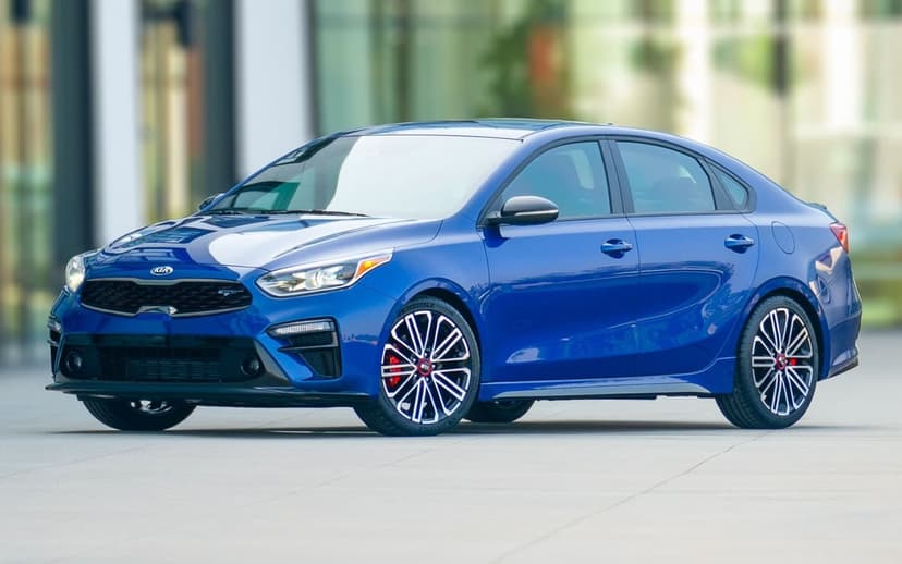Blue Kia Forte GT sedan with sporty alloy wheels parked in front of a blurred glass building.