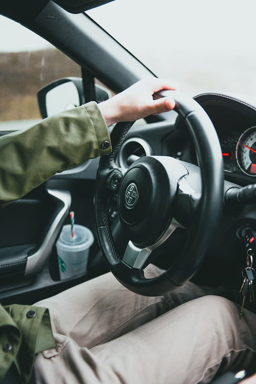 Hands grip a car steering wheel next to a coffee cup in a door pocket, viewed from the driver's perspective.