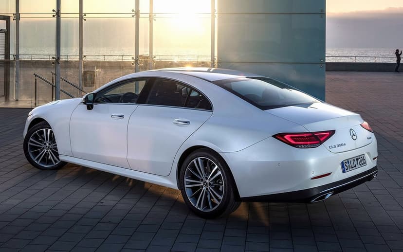 White Mercedes CLS 350d sedan parked on a tiled waterfront promenade against a glass building at sunset.