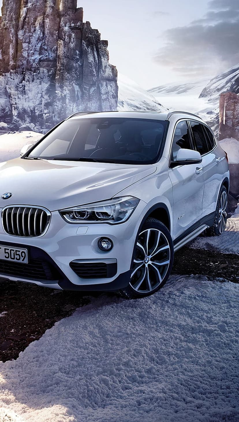 White BMW SUV driving along a winding road surrounded by steep, snow-covered cliffs and rocky mountain peaks.