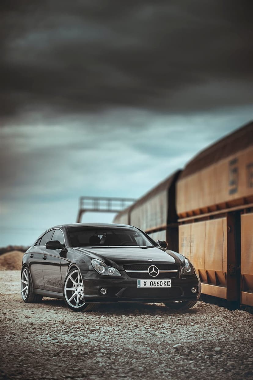 Black Mercedes CLS 350 sedan parked on gravel in front of weathered tan train cars under a dark overcast sky.