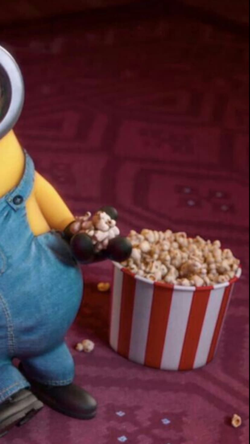 Yellow Minion in overalls holds popcorn next to a red striped bucket inside a dimly lit movie theater aisle.