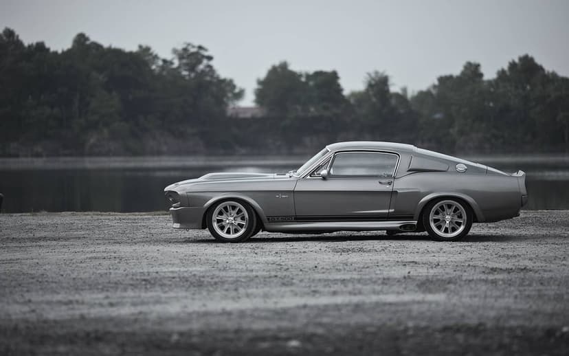 Silver Shelby Mustang GT500 Eleanor in profile on gravel with a blurred lake and forest background.