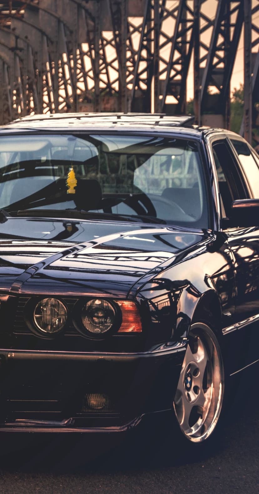 Black BMW E34 M5 with chrome wheels parked on a bridge during a golden sunset, featuring iconic dual headlights.