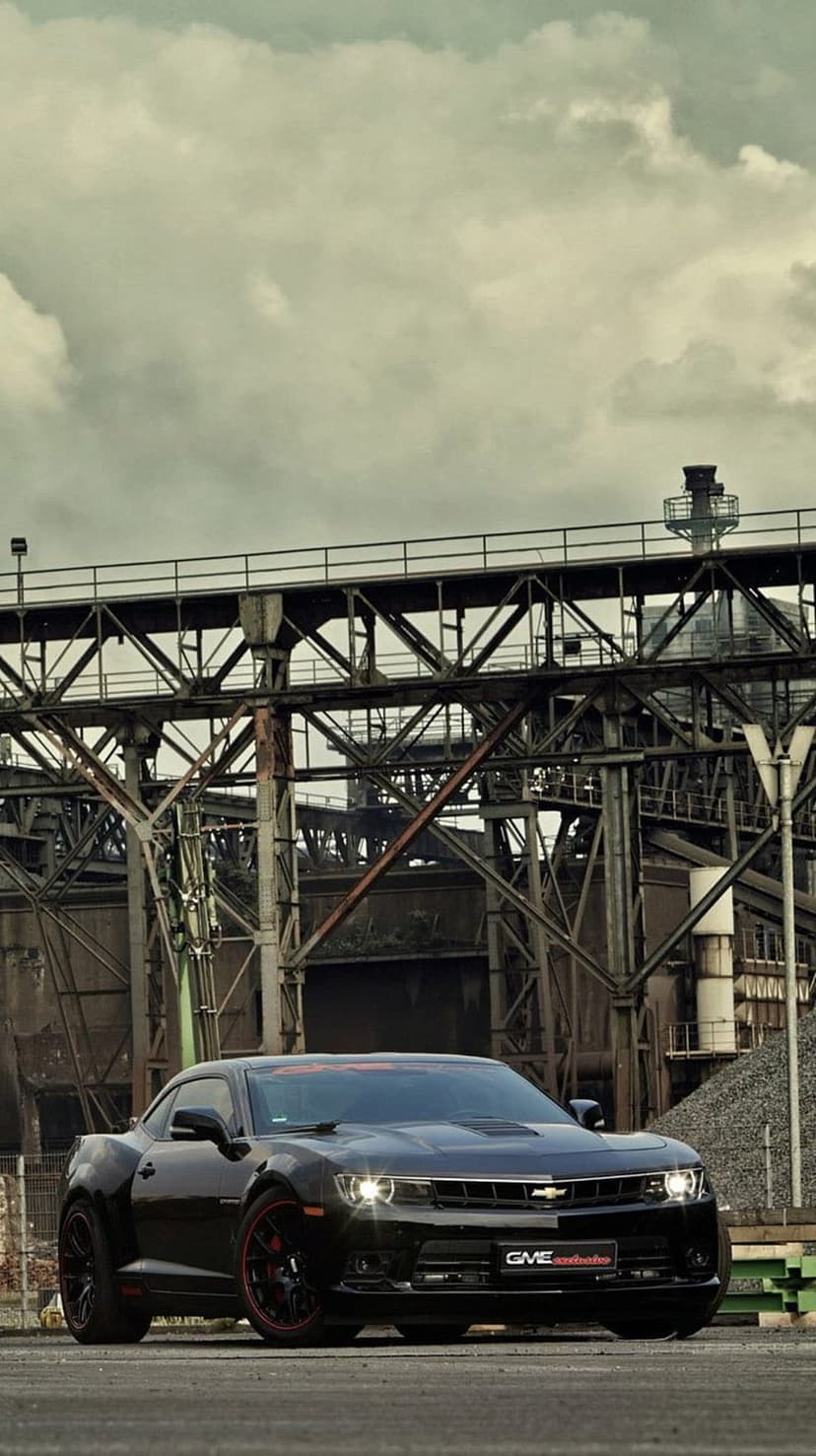 Black Chevrolet Camaro parked near rusted factory pipes under a dark cloudy sky in a vertical composition.