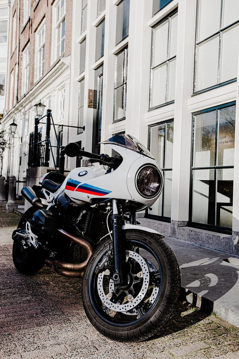 White BMW motorcycle with M-sport stripes parked on a cobblestone street in front of a historic stone building.