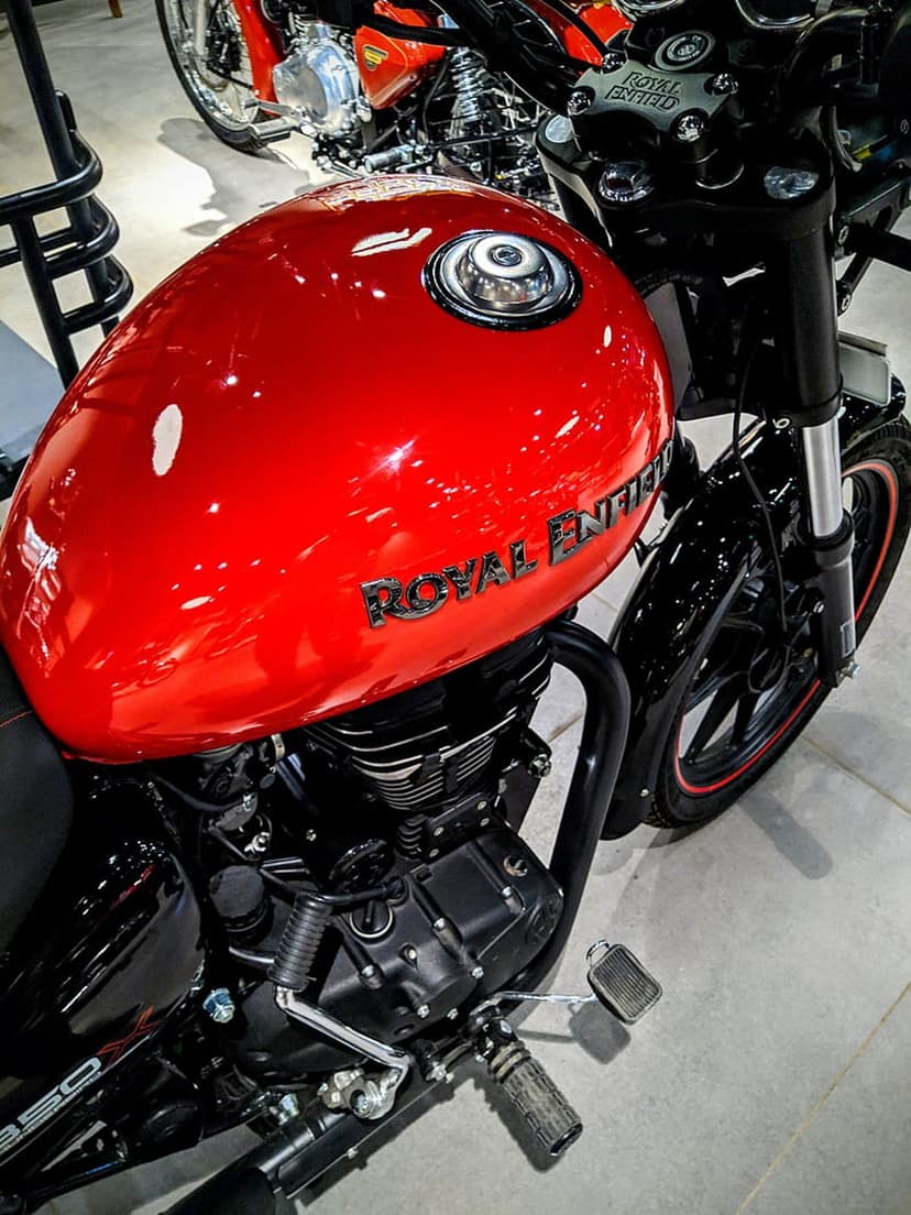 Red Royal Enfield motorcycle fuel tank with chrome badging, a metallic cap, and black engine components nearby.