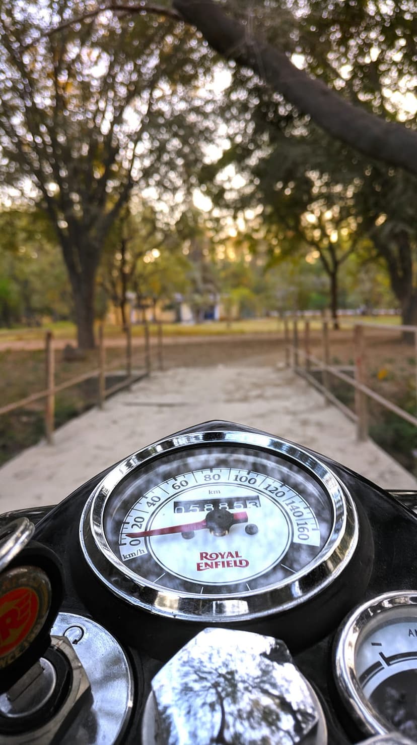 Royal Enfield speedometer and ignition key in focus against a blurred rural road with trees and a fence.