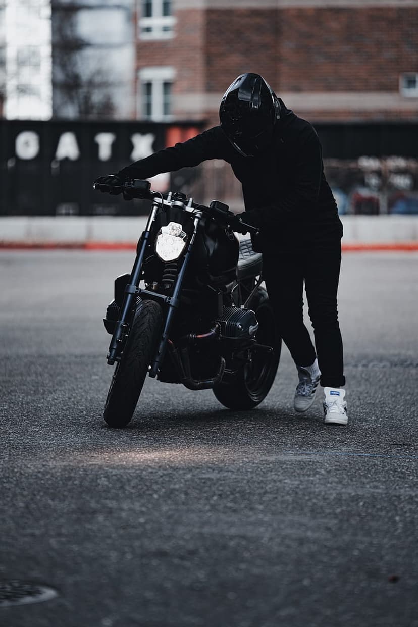 Biker in black gear leans on a custom motorcycle with a glowing headlight against a moody urban background.