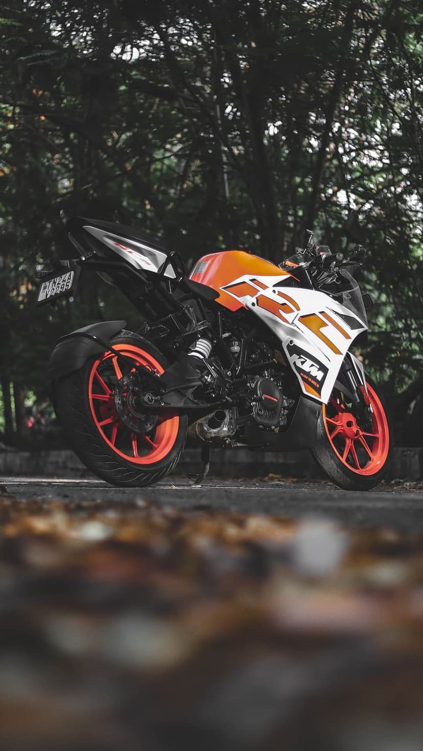 KTM RC motorcycle in orange and white parked on an asphalt road with fallen autumn leaves and green trees behind.