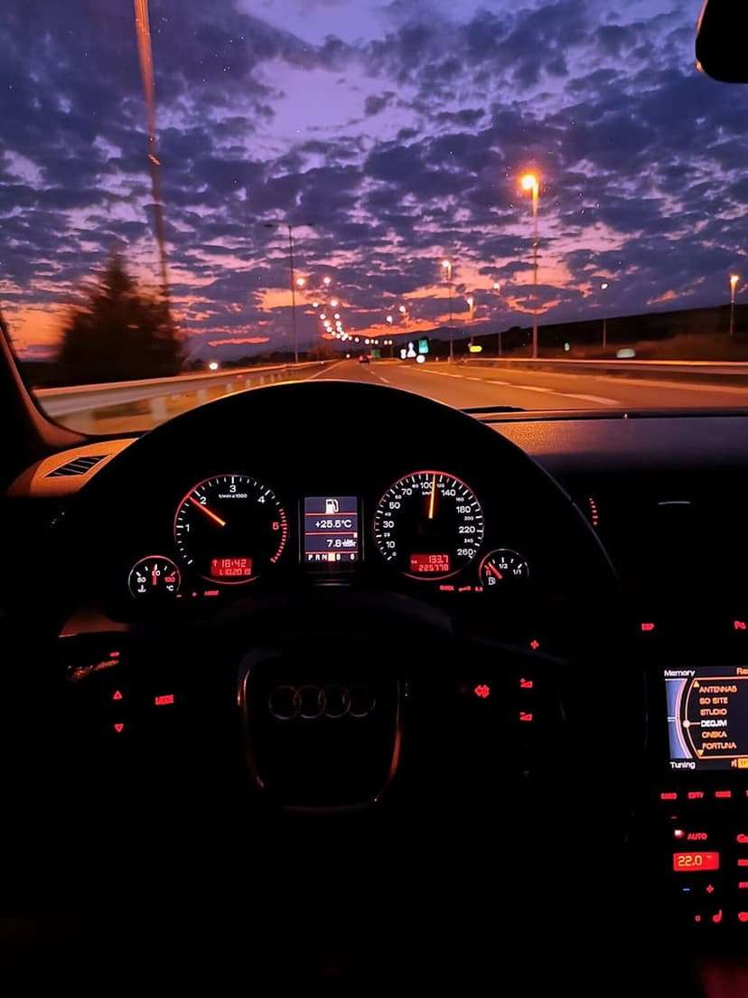 Red illuminated car dashboard and speedometer viewing a deep purple cloudy sky during an evening highway drive.