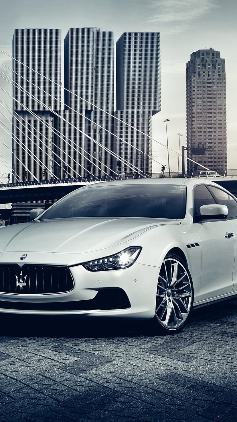 White Maserati Ghibli parked on cobblestones against a backdrop of tall glass skyscrapers in downtown Rotterdam.