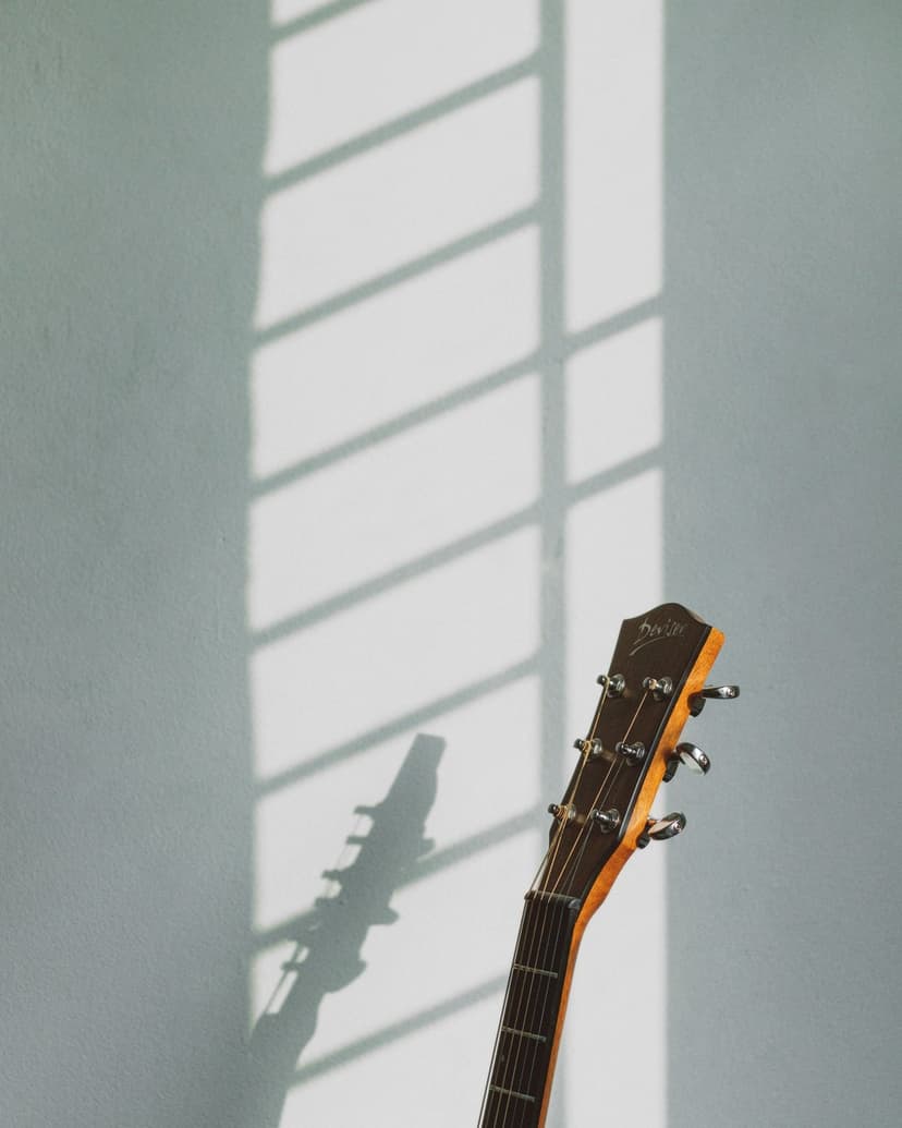 Acoustic guitar headstock and neck casting a lattice shadow on a pale blue wall under bright natural sunlight.