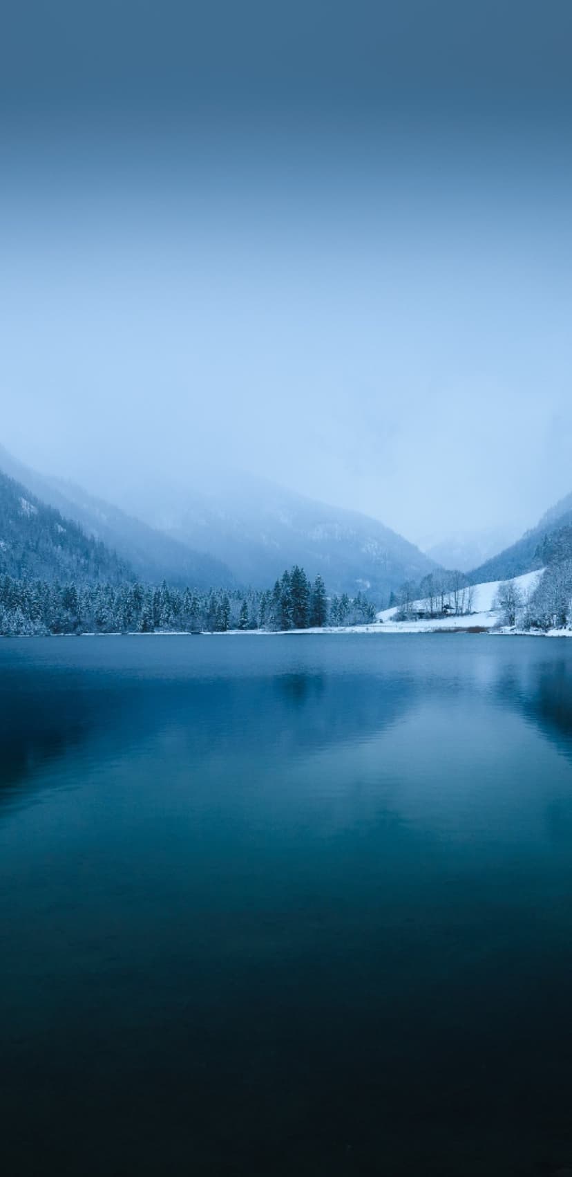 Blue mountain lake reflects snow-covered peaks and a misty evergreen forest under a soft, overcast winter sky.