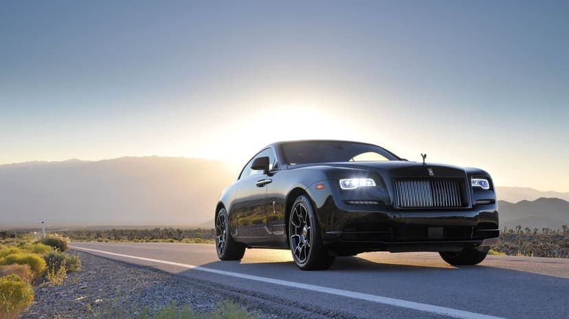 Black Rolls-Royce luxury car on a desert road with glowing headlights against a bright golden sunset.