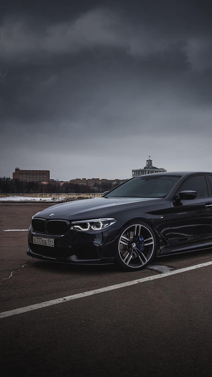Dark BMW M5 sedan with glowing headlights parked on a city street under a heavy, overcast stormy sky at dusk.