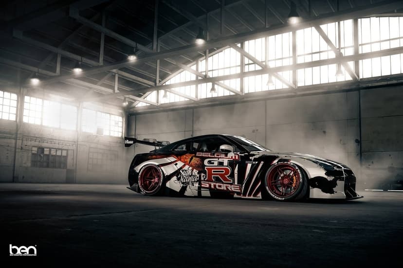 Nissan GT-R R35 with custom racing livery and red wheels in a hazy industrial warehouse with overhead lighting.