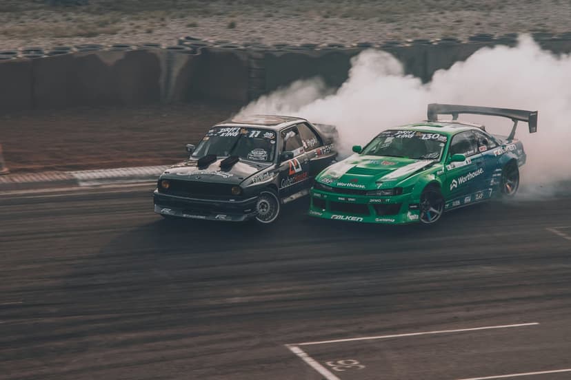 Black and green drift cars slide sideways on a track while emitting heavy white tire smoke in a vertical view.