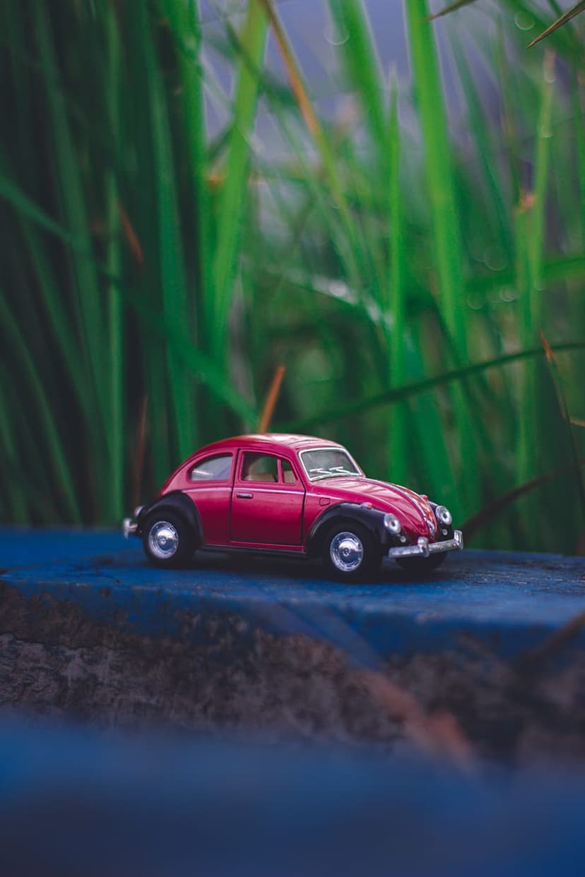 Red toy Volkswagen Beetle parked on a blue surface with blurred green grass and sky in the background.