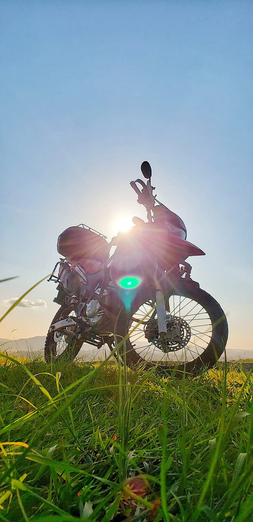 Motorcycle silhouette rests in tall green grass against a radiant orange sunset sky with a warm lens flare.
