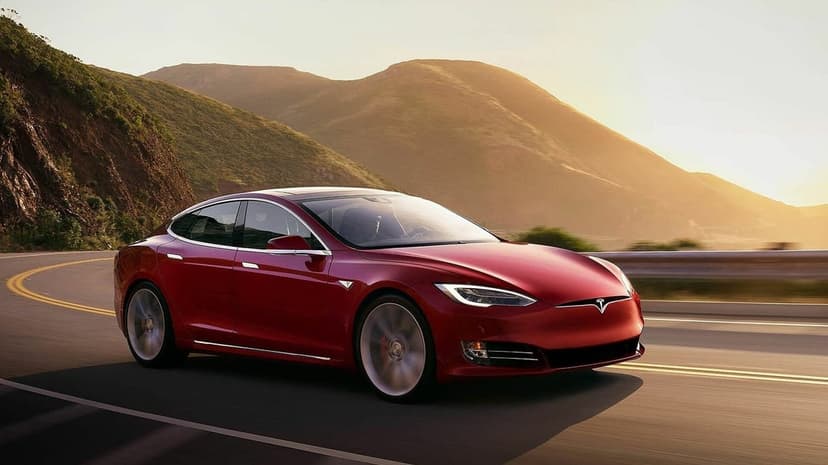 Red Tesla Model S driving on a winding coastal road at sunset with golden mountains in the background.