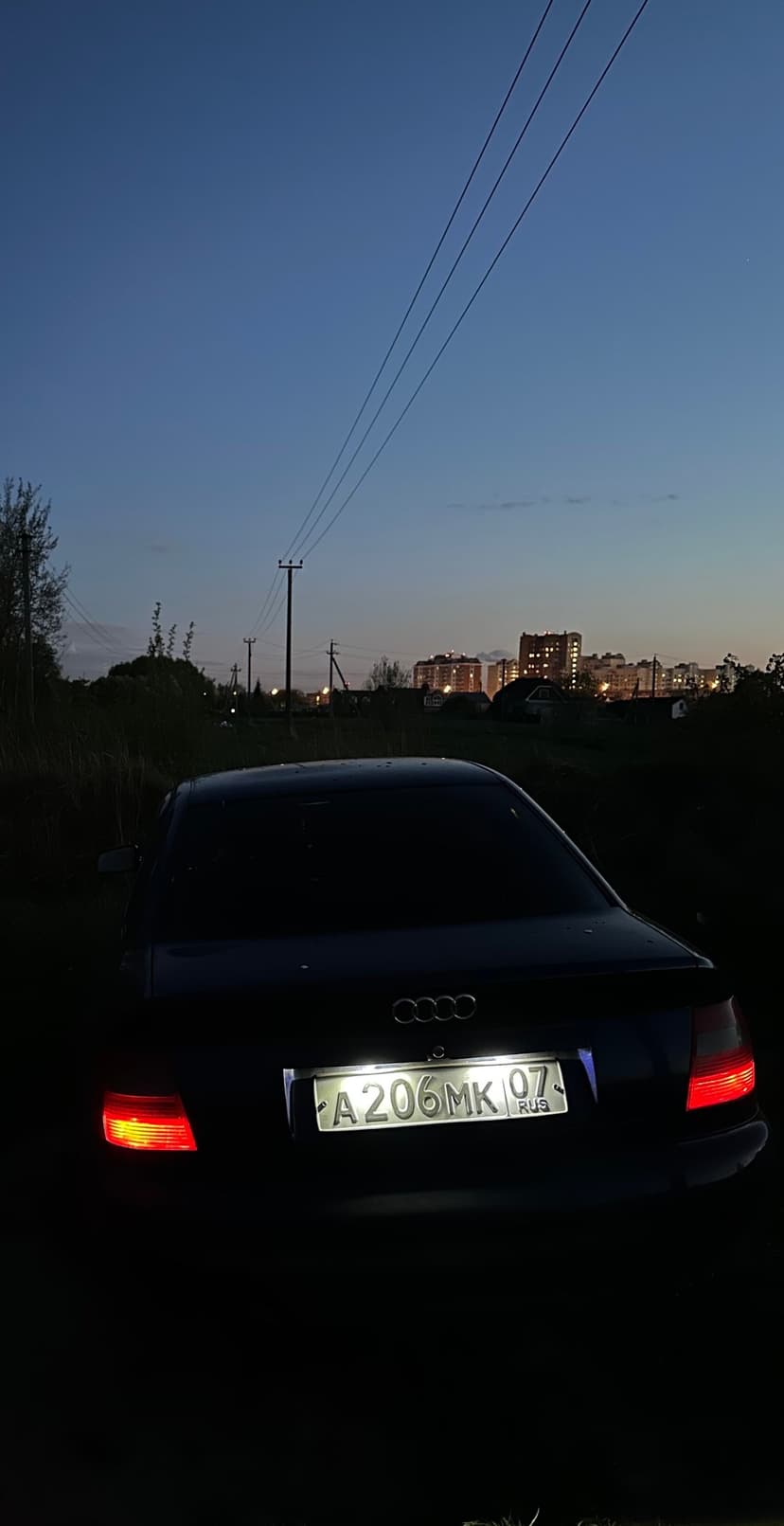 Dark Audi sedan with red taillights parked on a dirt road during twilight with a glowing city skyline background.