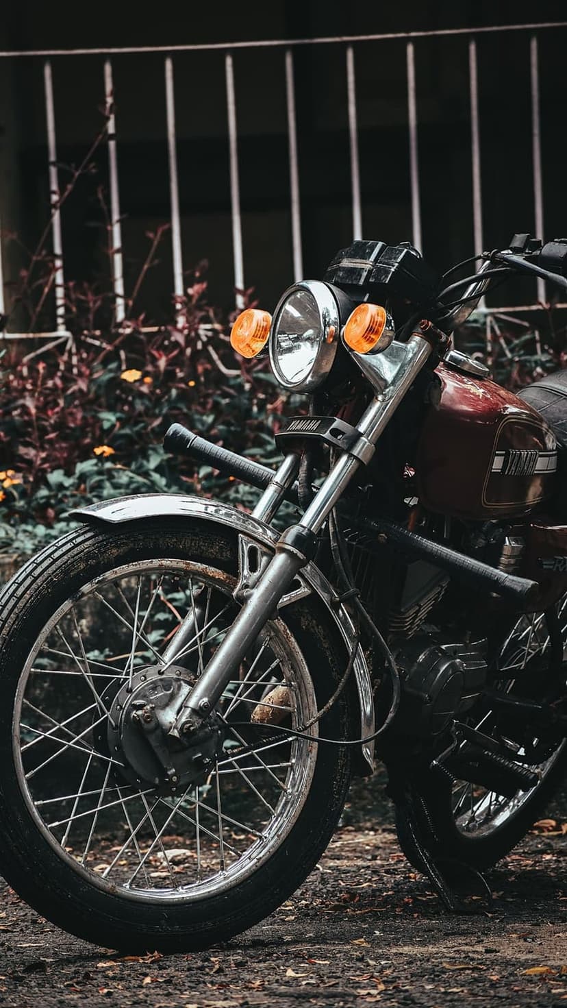 Red Yamaha motorcycle front wheel and chrome headlight against a white picket fence and green garden foliage.