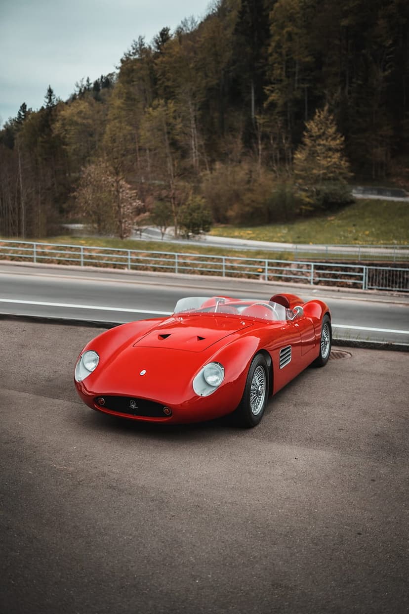 Vintage red Maserati race car with an open cockpit parked on a road beside a dense green forest under a blue sky.