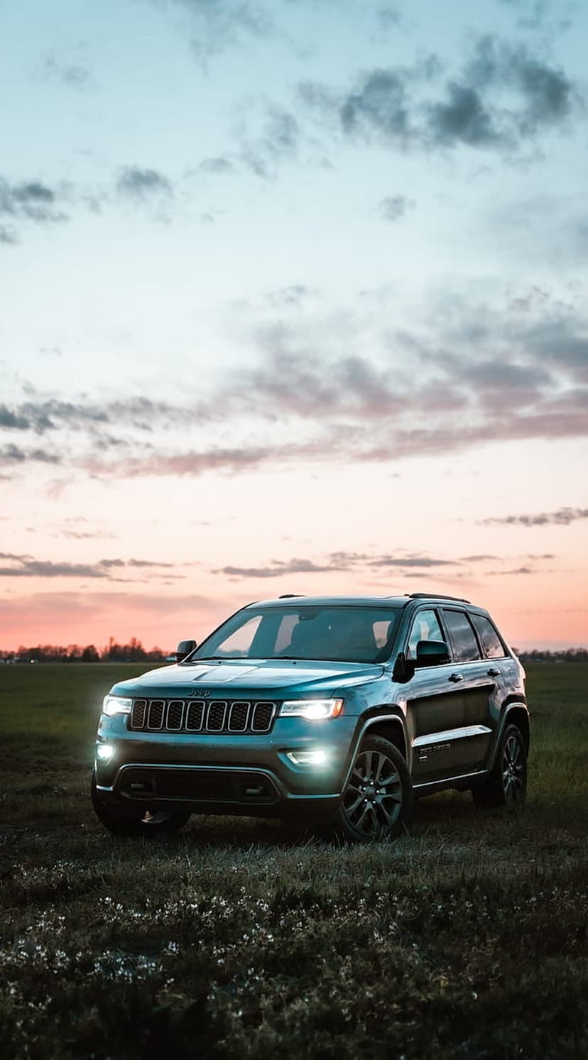 Jeep Grand Cherokee SUV parked in a grassy field with headlights on under a pink and orange sunset sky.