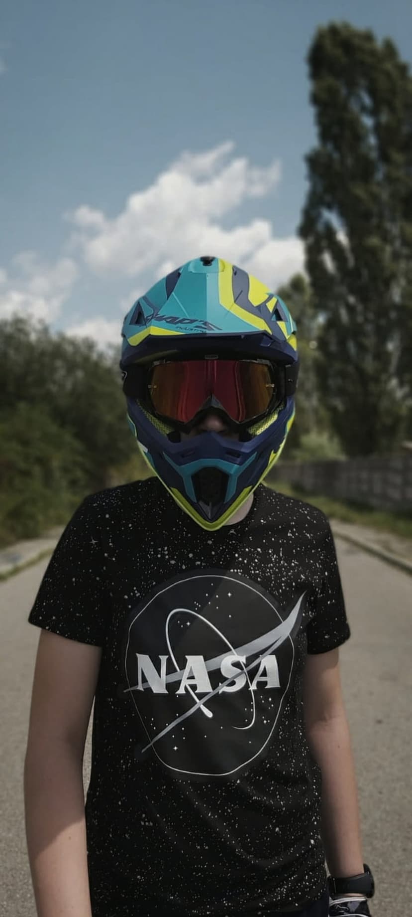 Person in a NASA t-shirt and racing helmet stands on an asphalt road with green trees and a blue sky behind them.